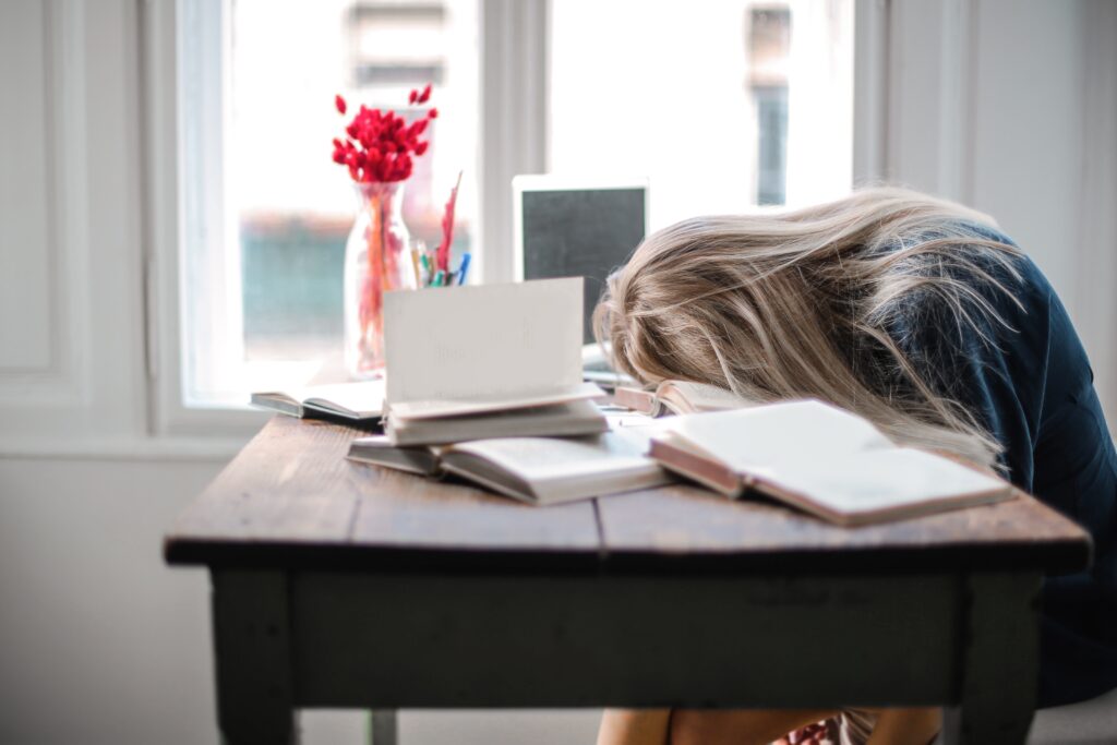 A picture of a girl putting her head down onto a wooden table as she she is being surrounded by books.