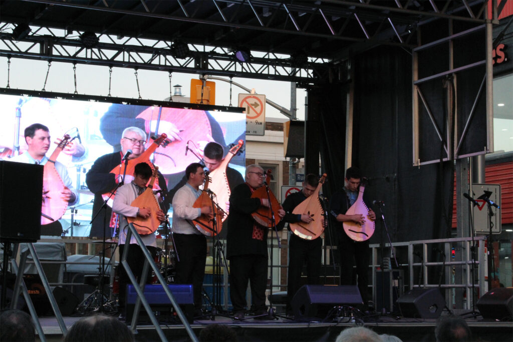 Five men are on stage holding lute like instruments. They preform in front of an audience. 