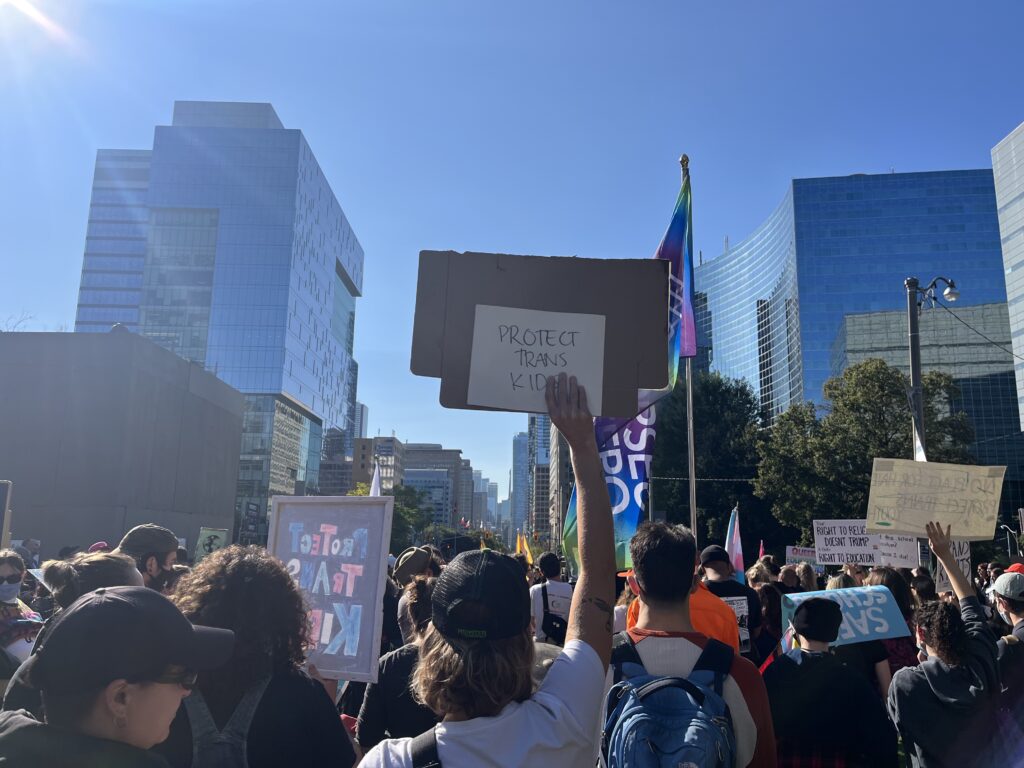 A group of pro-2SLGBTQ+ educational resources stands at Queen's Park in Toronto. In the foreground, someone is holding a sign reading "protect trans kids."