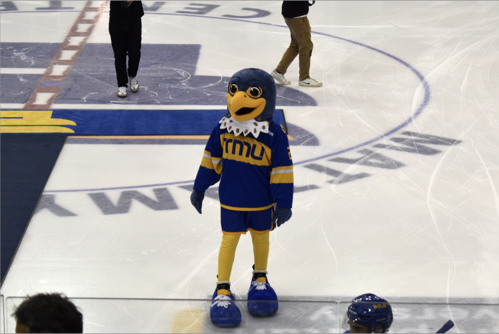 A photo of TMU falcon mascot Franke B. Bold in a blue and yellow falcon costume, standing on the ice of the Mattamy hockey arena.