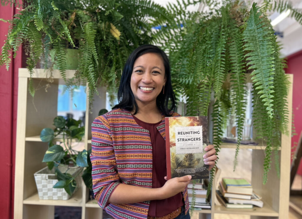 Author Jennilee Austria-Bonifacio stands in front of a bookshelf smiling and holding her novel, Reuniting with Strangers. Green plants sit on and in the bookshelf.