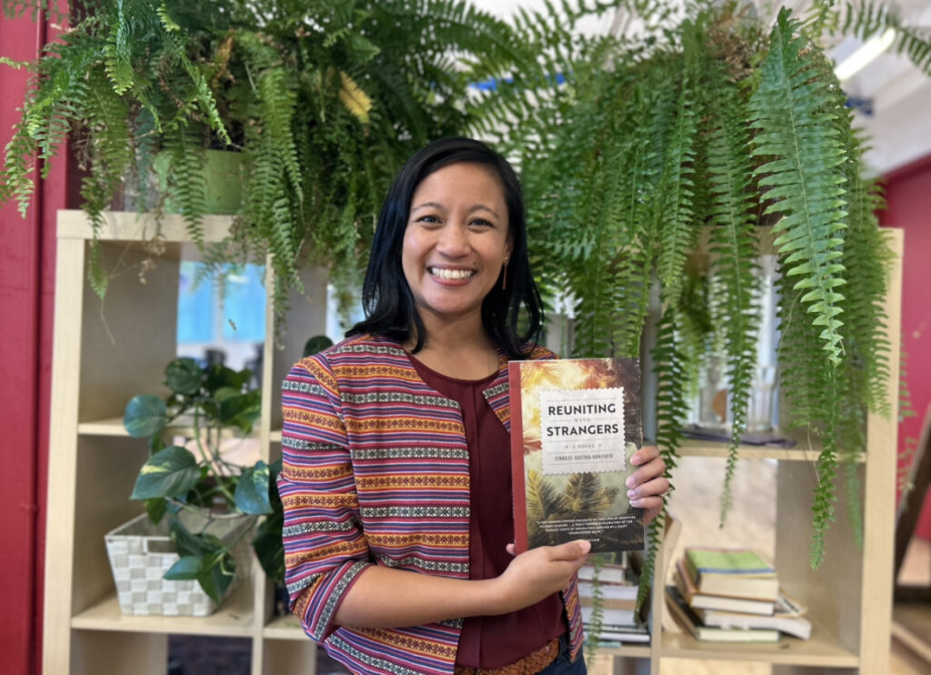 Author Jennilee Austria-Bonifacio stands in front of a bookshelf smiling and holding her novel, Reuniting with Strangers. Green plants sit on and in the bookshelf. 