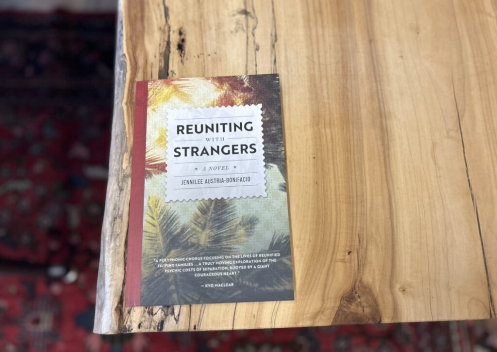 The novel Reuniting with Strangers by Jennilee Austria-Bonifacio is sitting on a wooden tabletop. In the background is a red carpet. 