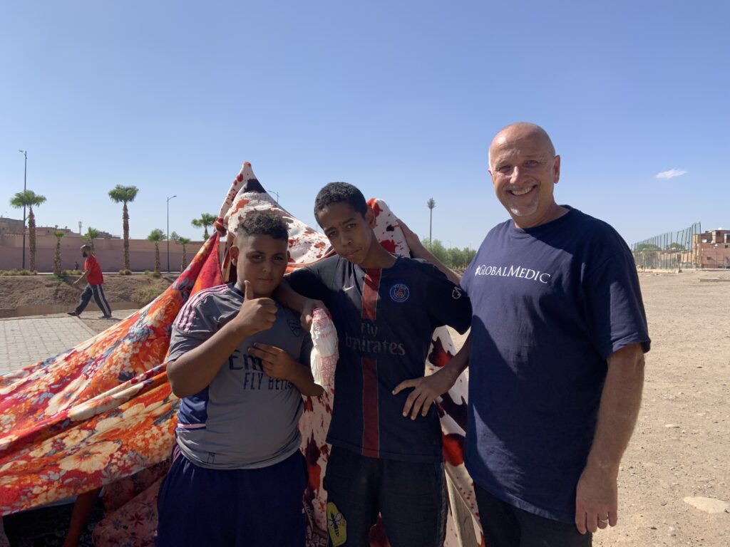 A member of the GlobalMedic response team stands, smiling, next to two young Moroccan boys, one of whom is giving the camera a thumbs up. They are all standing in front of what appears to be a makeshift tent.