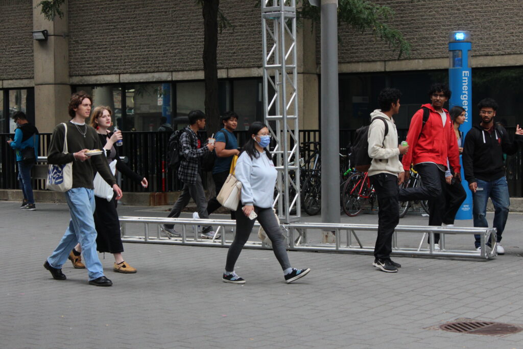 Students are seen walking down Gould St. on the TMU campus. The main subject of this image walks past wearing a blue medical mask.