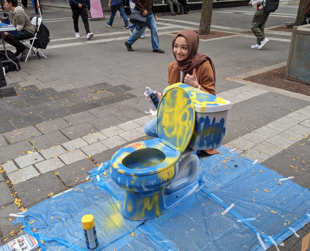 Student crouches while posing next to a toiler that has been spray painted blue and yellow.