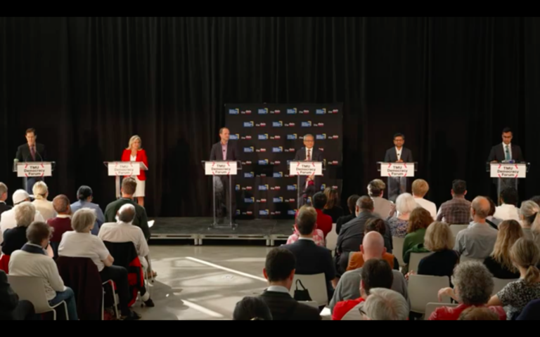 Debate moderator stands on the dais with five Liberal Party of Ontario candidates.