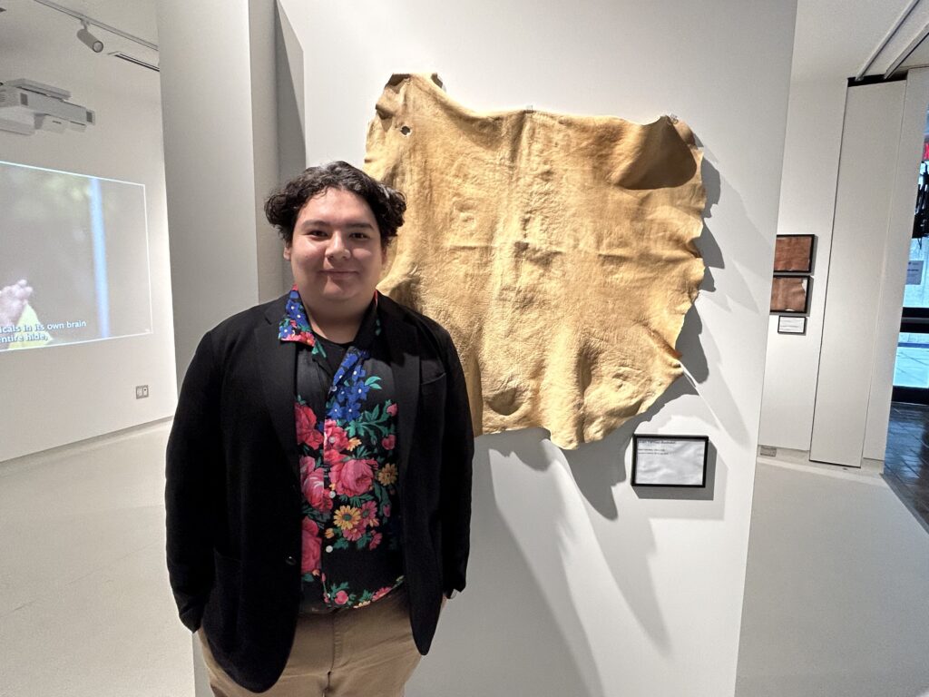 Caleb Wesley (a person wearing a black blazer over a flowered shirt with tan pants) poses next to a sample of tanned deer hide.
