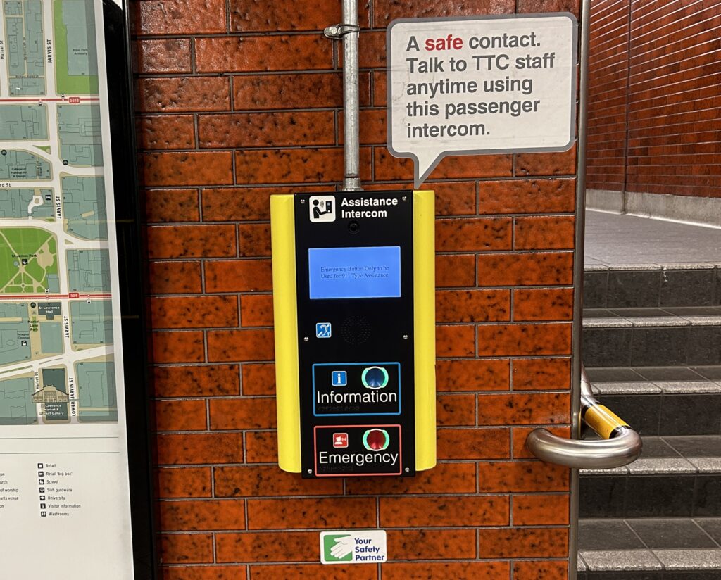 An emergency assistant button (a yellow box with several buttons for information and emergency) is shown at the bottom of the stairs of Queen Station's entrance