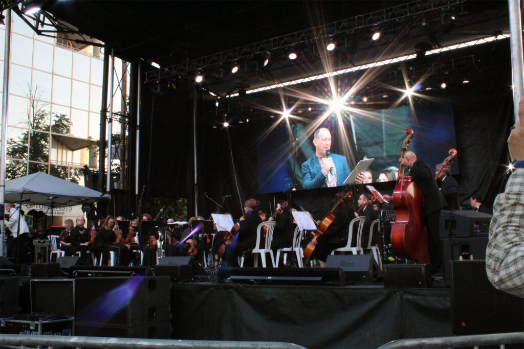An orchestra preforms on stage with multiple instruments. A man stands on the side of the stage with a microphone.