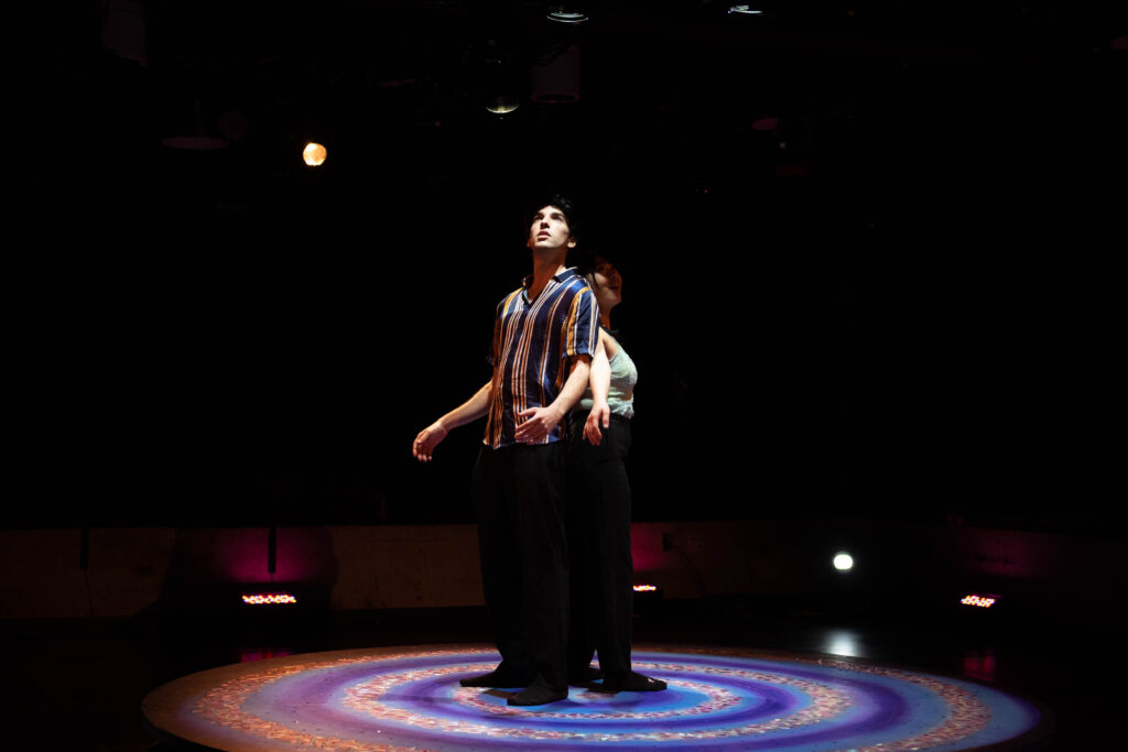 Two people stand close together back-to-back on stage under a blue concentric spotlight. The man is in front of the woman, casting his shadow on her.
