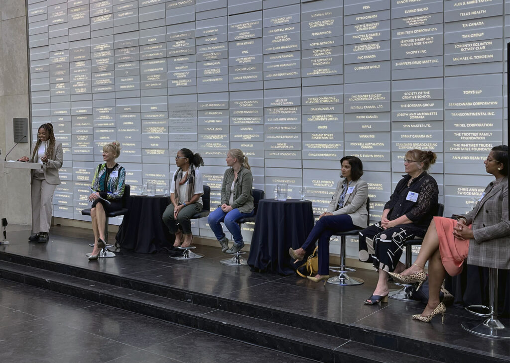 Seven women on a stage in a brightly lit room. One Woman stands behind a podium.