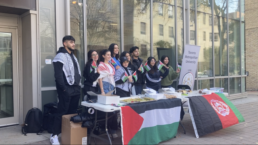 students standing outside at a table with baked goods, a Palestinian flag and an Afghan flag.