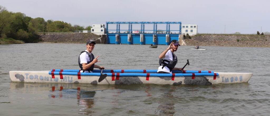 Two members of Concrete Canoe sit in the team's 2023 canoe on open water, showing the boat's cow print design,