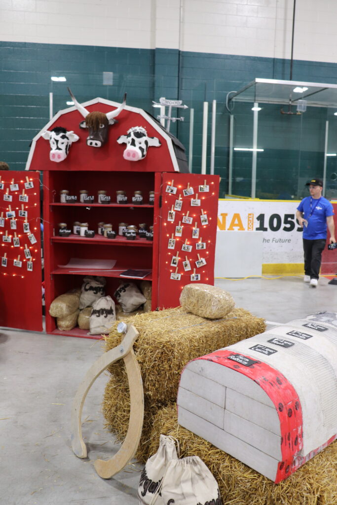To enhance their 2023 cow theme, T-Moo, the team displayed their concrete mix ingredients in a small, red barn, with string lights and polaroids. The barn has three cow heads on top and in the foreground are three hay bales.