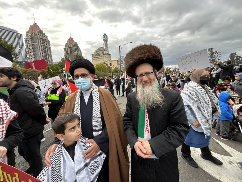 (From left to right) Islamic cleric places his hands on his sons' shoulders as he stand next to an Hasidic Ultra-Orthodox Jewish man. The cleric and his son are both wearing the traditional Palestinian scarf on their necks, while the Ultra-Orthodox Jew is wearing a scarf donning the colours of the flag of Palestine with a red inscription "PALESTINE" at a pro-Palestinian rally at Mississauga, ON, against Israeli bombardment and siege of the Gaza Strip. Other protestors and buildings are seen in the background.