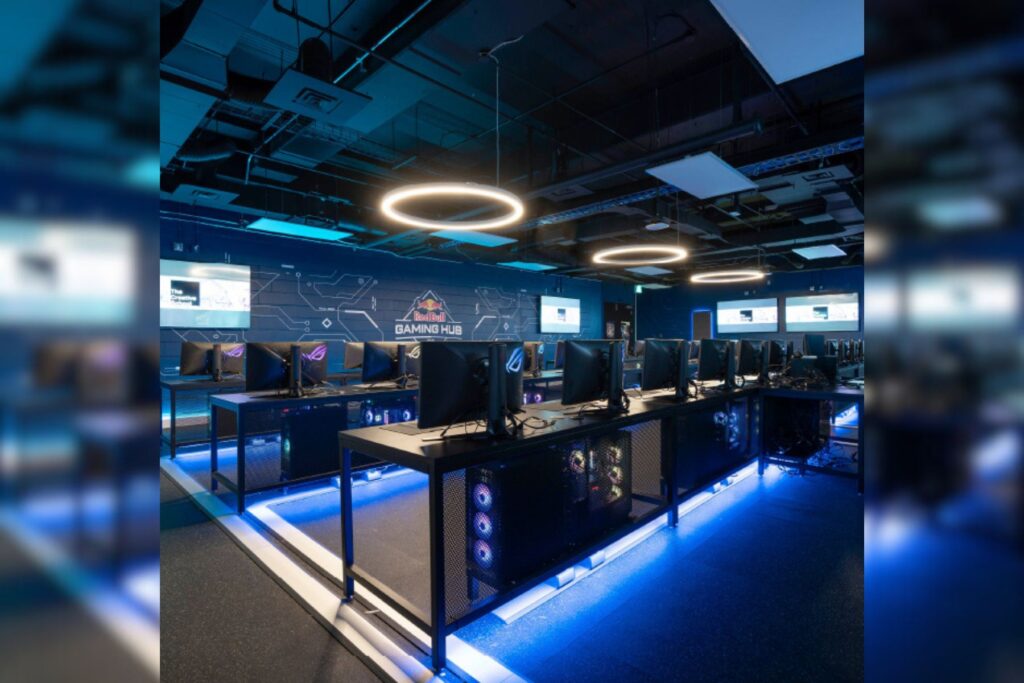 The Red Bull Gaming Hub at TMU features a room with rows of long desks, lined with individual gaming stations. Each station features a monitor, keyboard and mouse, as well as a 'gaming chair,' similar to an office chair. Blue LED lights shine from under each row of tables, and the back wall features an ornate electronic wire-like pattern with the RedBull logo in the middle.
