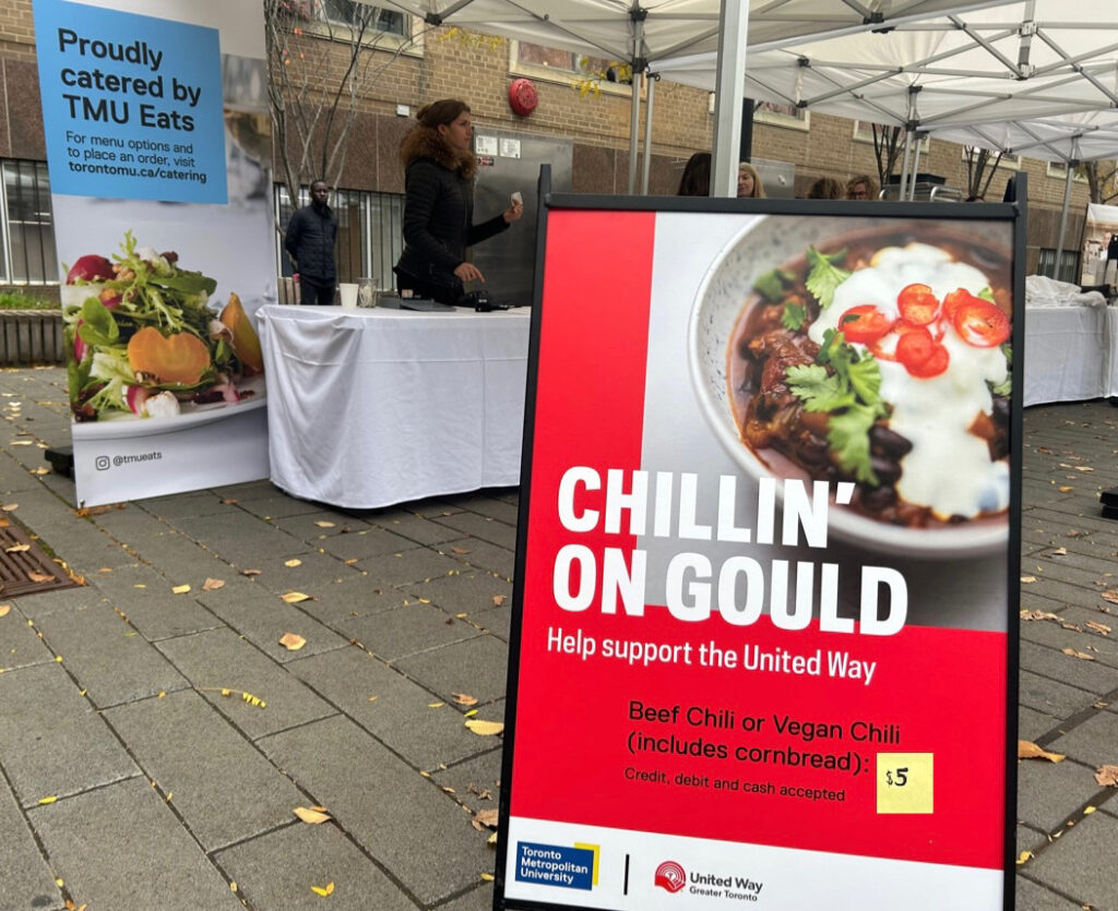 A red sign with white text reads "CHILLIN' ON GOULD. Help support the United Way." In the background is tables set up, and another sign that reads, "Proudly catered by TMU Eats."
