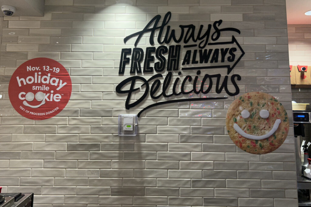 A wall with the black text "Always fresh, always delicious." To the right is a red sticker with the text "Nov. 13-19, Holiday smile cookie" and an image of the cookie is to the right.