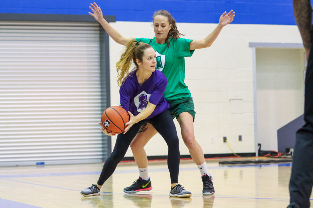 Player in purple holds basketball while player in green blocks them