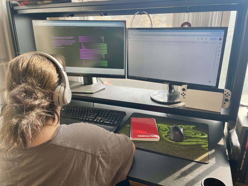 A woman sits at a desk. In front of her are two computer monitors, one with a blank google doc, the other on OpenAI's website, with text that reads: "Introducing ChatGPT". on the desk sits a red notebook with the word  "CANCULTURE" on it's cover.