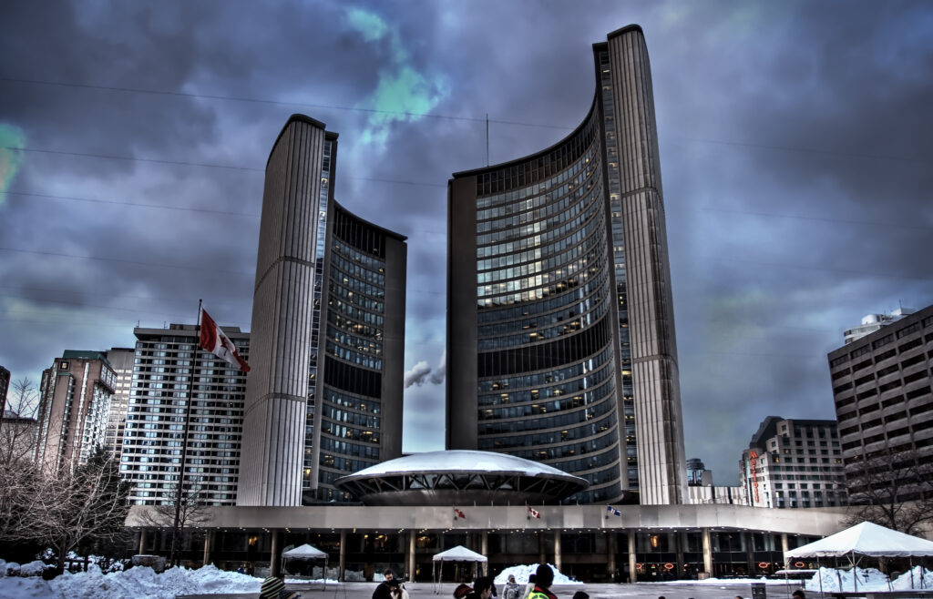 A photo of Toronto's City Hall building with snow on the ground with a cloudy sky and a Canadian flag waving on the left.