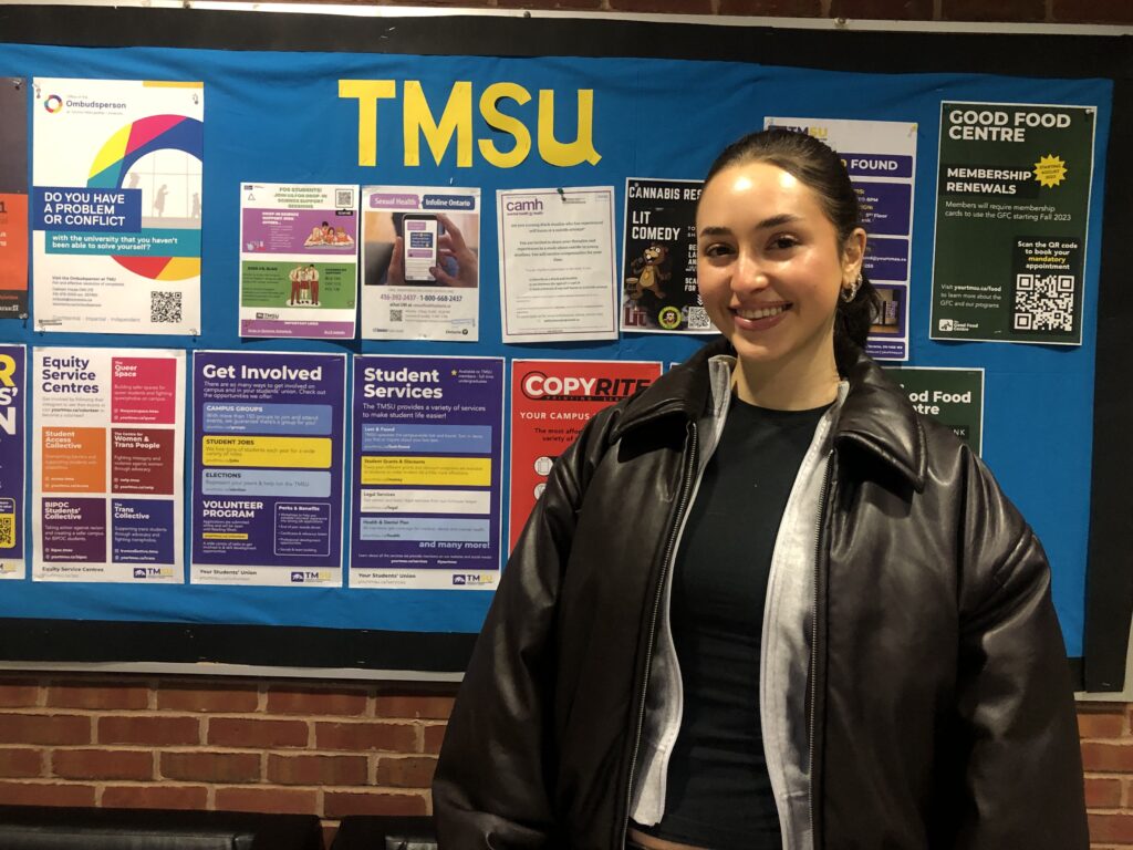 A woman wearing a black leather jacket smiles in front of a notice board with flyers from the Toronto Metropolitan Students' Union