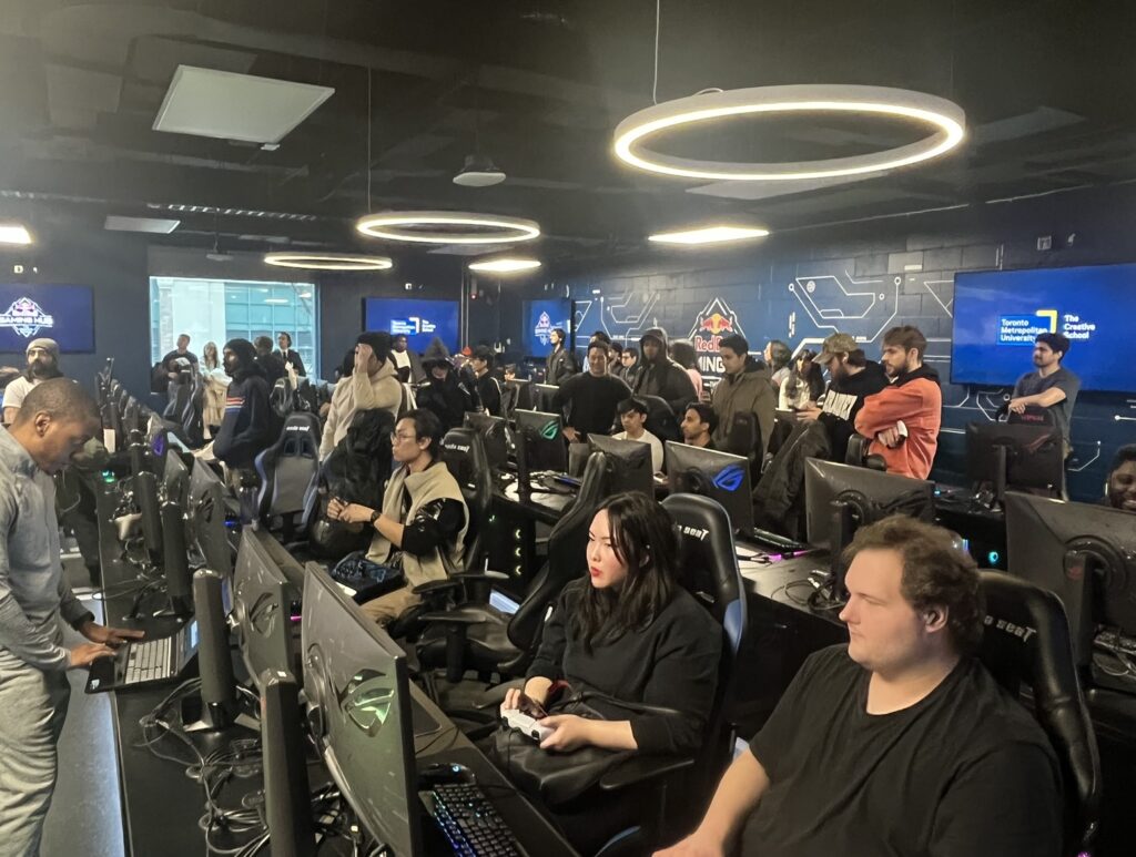 Room full of gaming computers, with a large group of people playing on them and more people standing on the sides watching their games