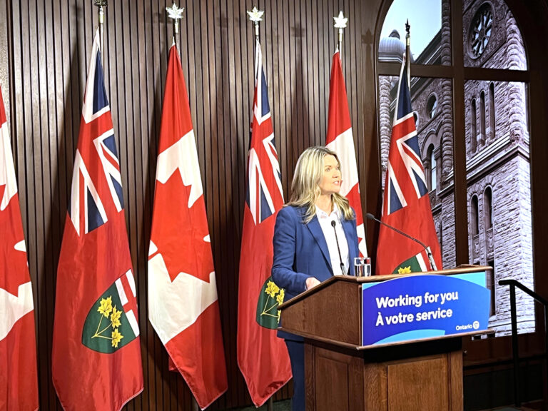 A blonde woman in a suit--Minister Jill Dunlop--stands at a podium which has the words "Working for you" written in English and French. Behind her are various Canadian and Ontario flags as well as a large picture of Queen's Park