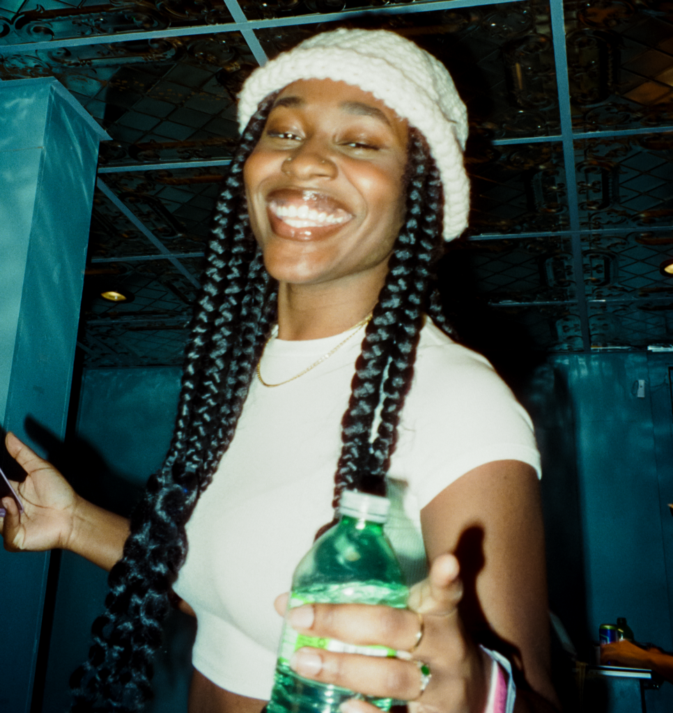 A woman in a small white hat with a white t-shirt holds a water bottle while smiling at the camera.