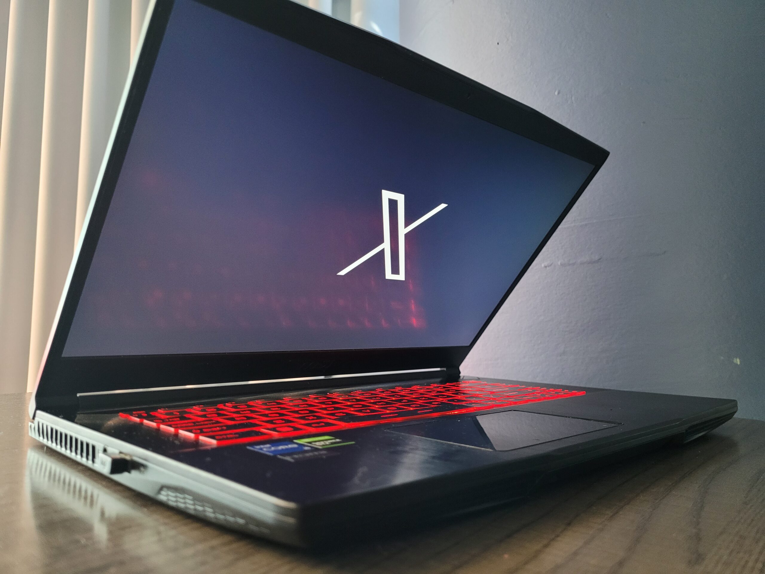 A half closed laptop with red keyboard lights in a dark room displays the X logo
