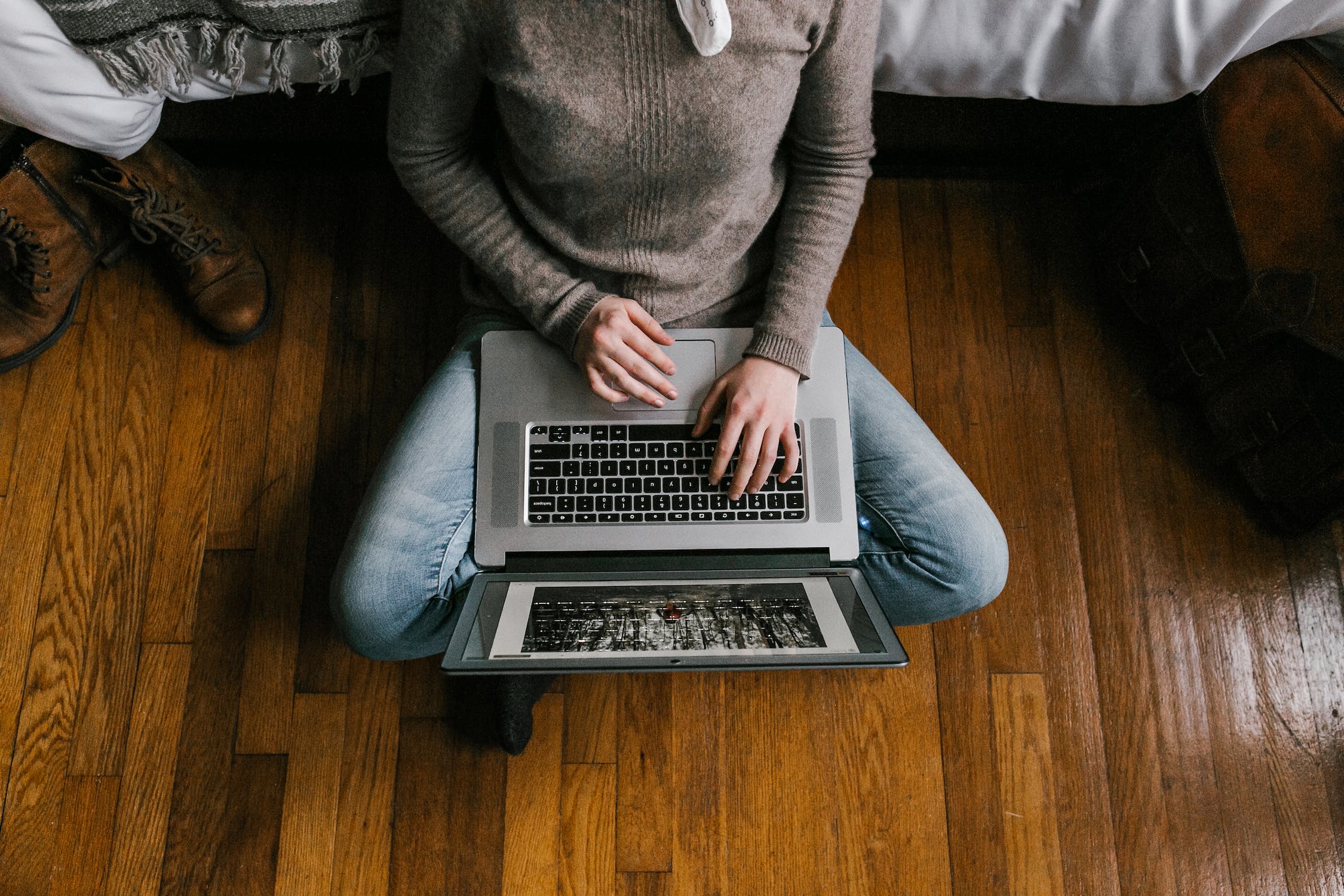 A person in blue jeans and grey sweater sitting on their floor against a bed with their legs crossed whilst scrolling through their computer.