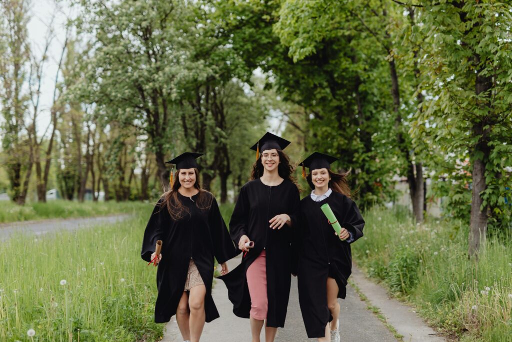 Three women dressed in black graduation caps and gowns walk happily down a trail surrounded by trees.