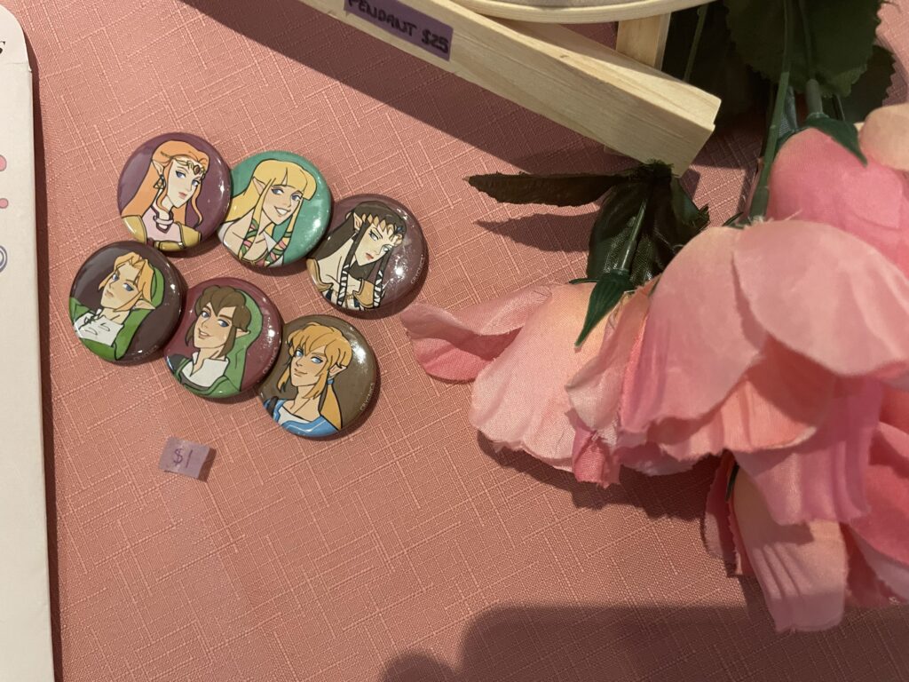 Six pins of cartoon-style portraits lay for sale on a table beside pink flower and a sticker that reads, "$1". 
