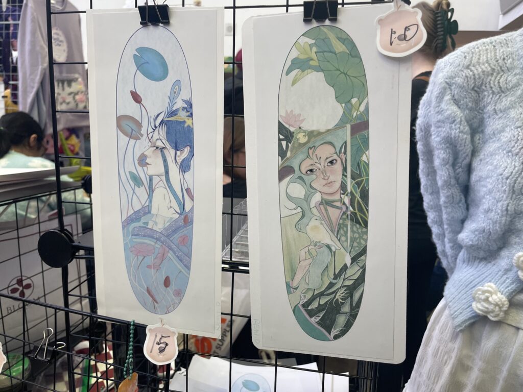 Two drawn portraits of a woman in green and blue hang for sale on a wired rack.