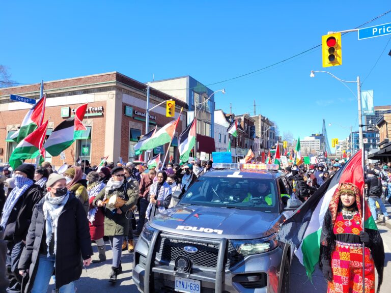 A crowd of demonstrators march down Yonge St., many holding Palestinian flags and wearing keffiyehs as they walk past a police vehicle.