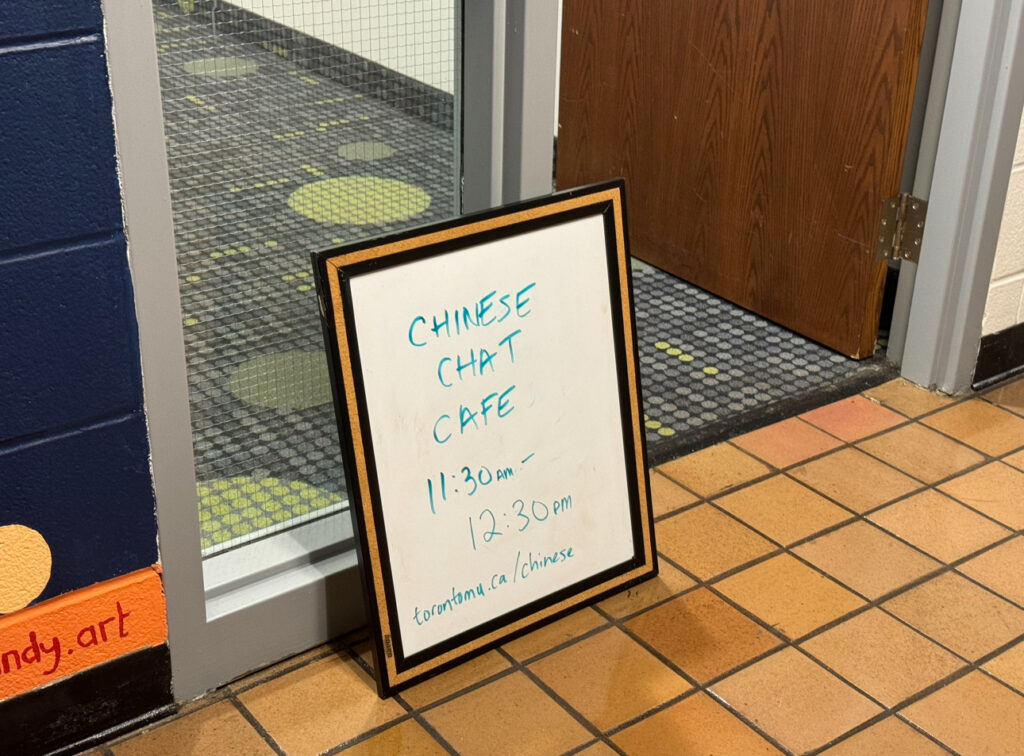 White board next to a door. White board has written "Chinese Chat Cafe 11:30 am-12:30pm"