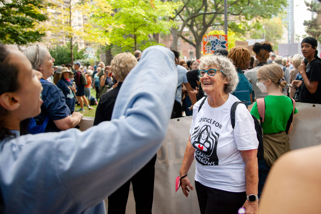 Rebick wears a white shirt reading "justice for Grassy Narrows" while smiling in front of a crowd of demonstrators 