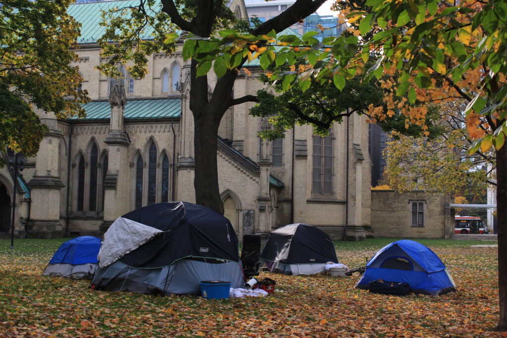 Four tents in a homeless encampment against the background of a church and trees, with a TTC bus visible in the background