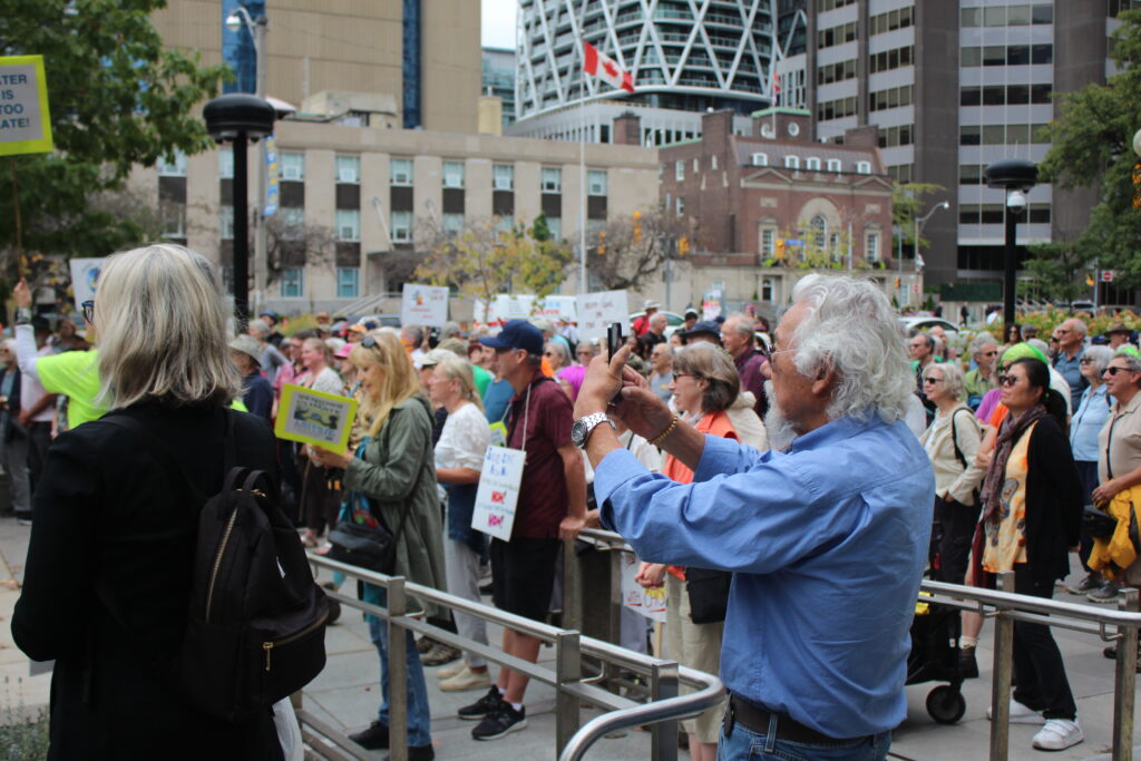 David Suzuki stands alongside a crowd, holding up his phone to take pictures.