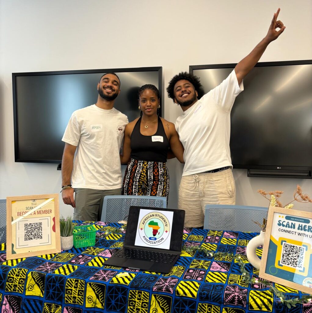 Three Black students stand in front of a table.