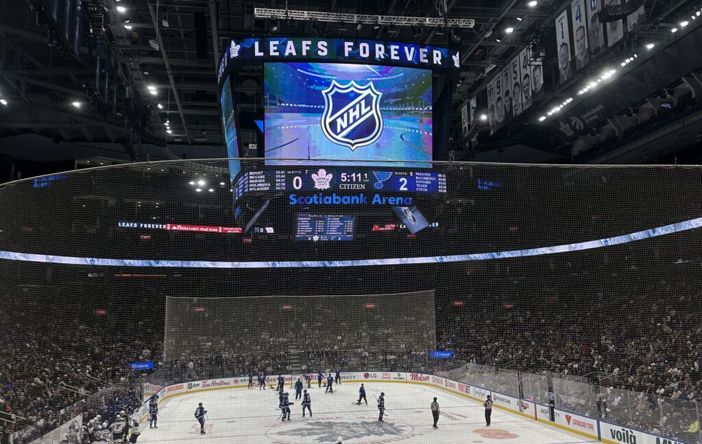 A photo of an arena with an ice rink, an audience of fans, and hockey players scattered on it. The large TV in the centre reads Leafs Forever.