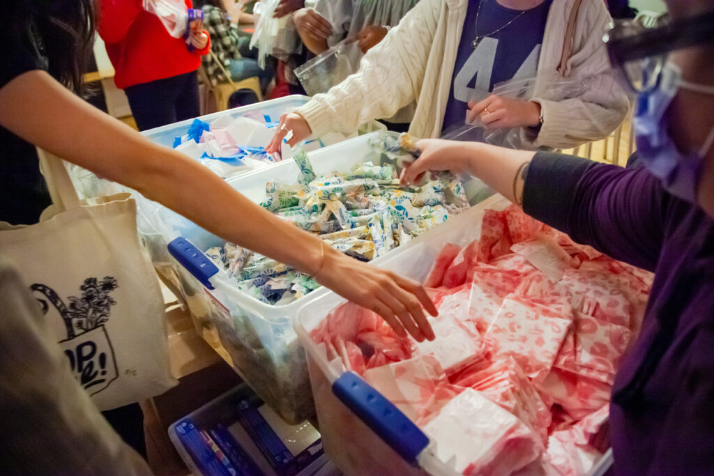 Three buckets containing period care products with arms reaching over each other and packing them into bags.