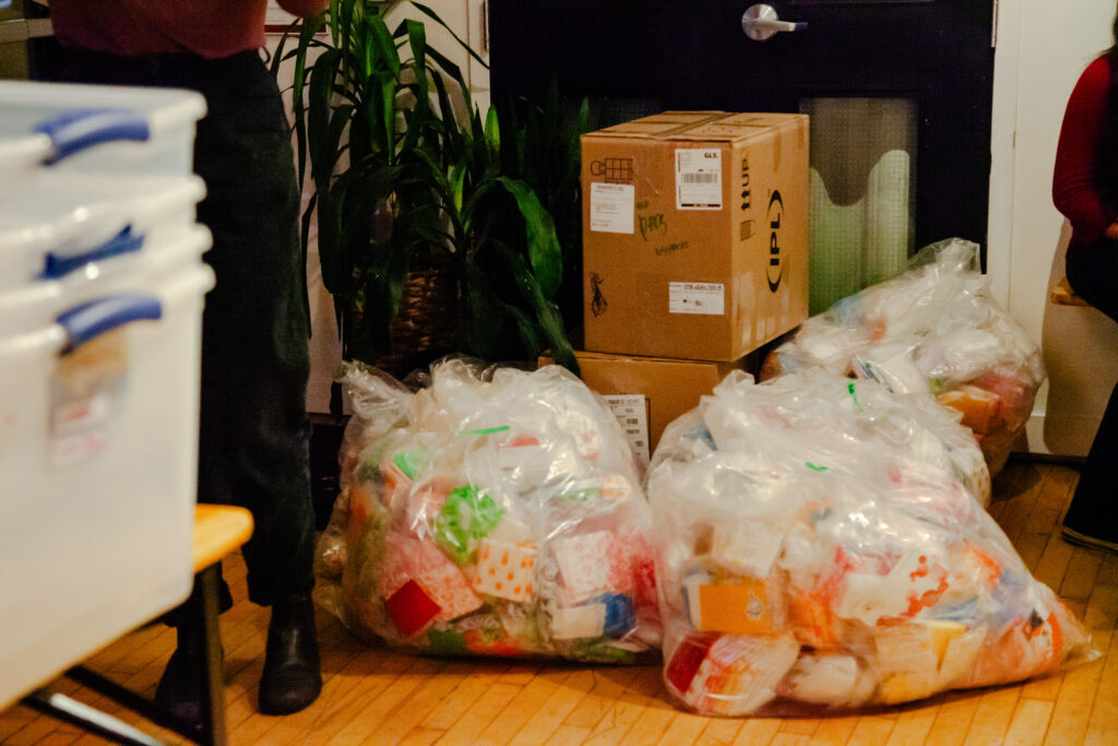 Three large plastic bags filled with period packs. A stack of boxes also containing the packs sit behind them. 