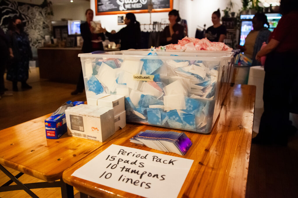 A table with buckets filled with period products. A paper stuck to the table reads "Period Pack. 15 pads. 10 tampons. 10 liners. A box of zipper seal bags sits next to it. 