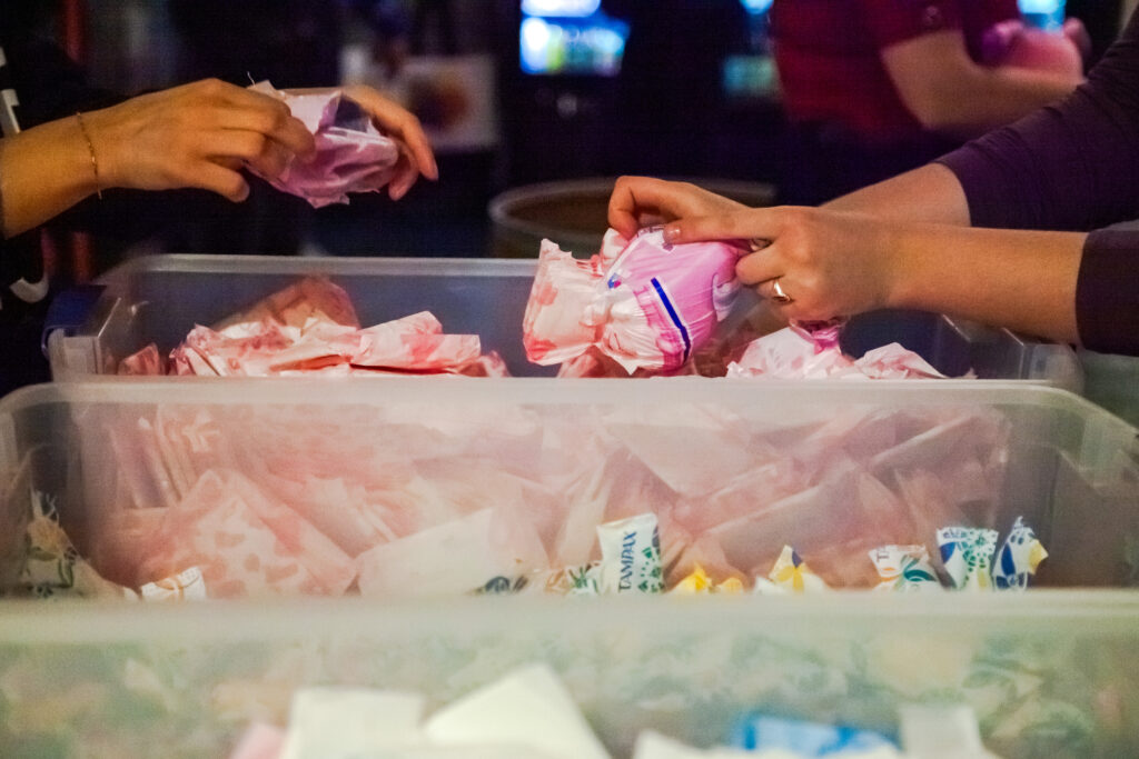 Three buckets  on a table filled with period products. A person refills the bucket by opening a pack of pads and emptying the individual products. 