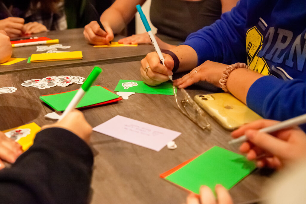 A group of people  write personalized messages on colourful cards. Some put stickers on the cards. 