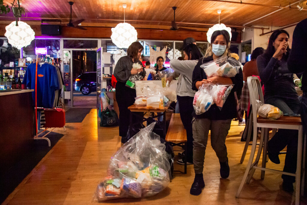 A person juggles four bags packed with period products. Around them, a scene of people mingling and assembling surrounds them.