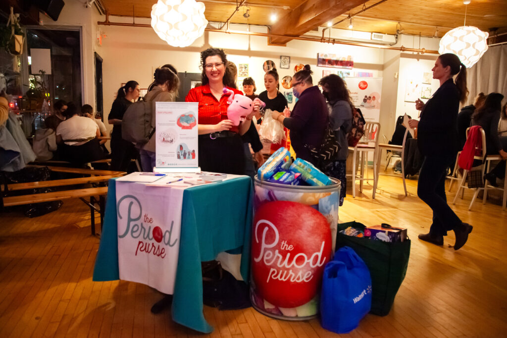 VanSickle smiles while holding a smiling uterus plushie behind The Period Purse info booth and donation bucket. The bucket is overflowing with donated products.