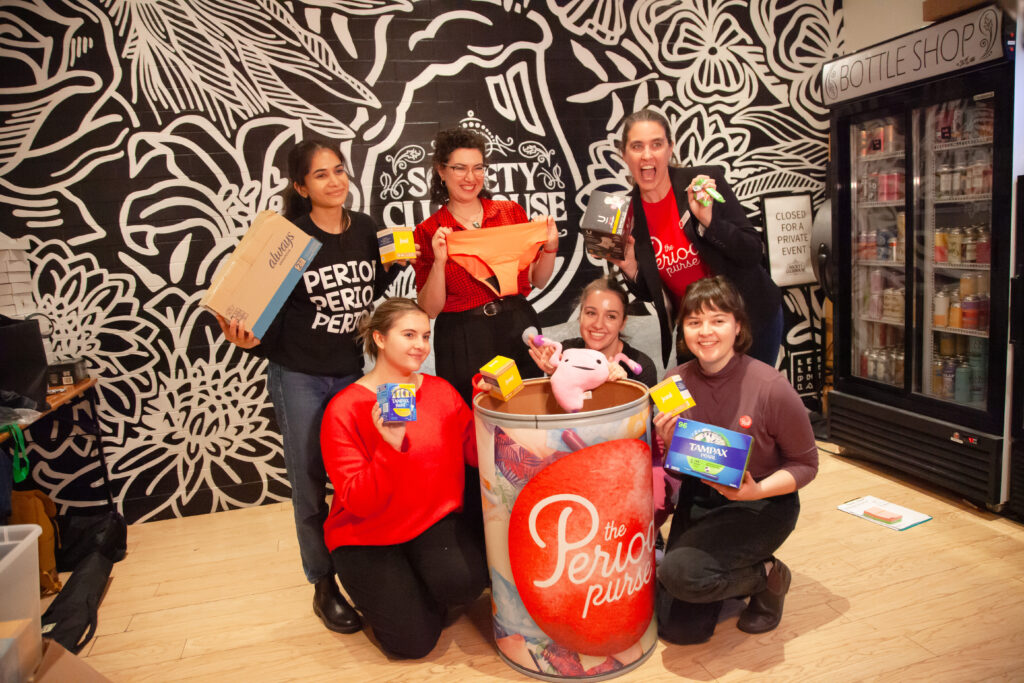 The Period Purse team pose for a photo behind a donation bucket with their logo. Everyone is smiling while holding varying menstrual care products.